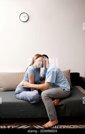 Una coppia si siede a stretto contatto su un divano nel soggiorno. Si sorridono mentre condividono un momento privato. L'orologio mostra l'ora serale. Foto Stock