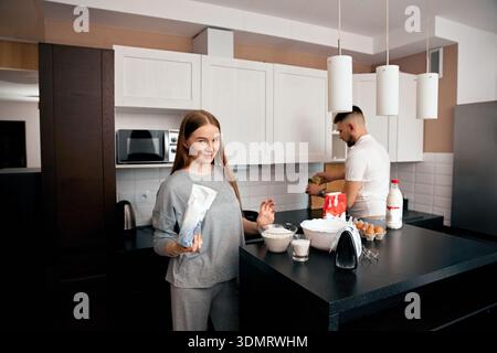 Una donna si trova sull'isola della cucina tenendo in mano una borsa mentre il suo partner prepara il cibo al bancone. Stanno lavorando insieme nella loro cucina di casa. Foto Stock