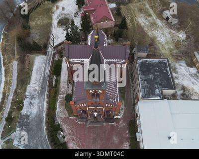 Vista aerea di una chiesa cattolica in mattoni rossi con architettura gotica, guglie alte e croce, circondata da neve, alberi, e negli edifici vicini. Foto Stock