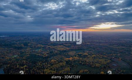 Vista aerea di un paesaggio lituano durante il tramonto, con foreste autunnali, campi aperti, un piccolo specchio d'acqua e un suggestivo cielo nuvoloso. Foto Stock