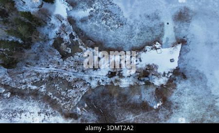 Una penisola innevata con una piccola cabina, alberi sparsi e sentieri visibili circondati da acqua ghiacciata, catturati dall'alto in Lituania. Foto Stock