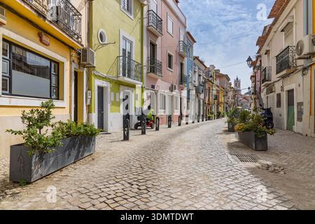 Affascinante strada acciottolata a Setubal, Portogallo, caratterizzata da case tradizionali colorate, balconi decorativi in ferro e piantagioni di pietra sotto un sk blu Foto Stock