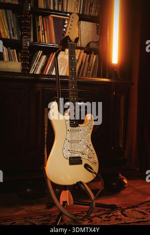 Una chitarra elettrica color crema poggia su un piedistallo in una stanza accogliente con una libreria e un'illuminazione calda. La scena mette in risalto l'atmosfera musicale e lo charac vintage Foto Stock