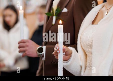 Un primo piano di una sposa e di uno sposo che tiene alte candele bianche durante la cerimonia nuziale, circondato da una luce soffusa e da accenti floreali in un chu Foto Stock