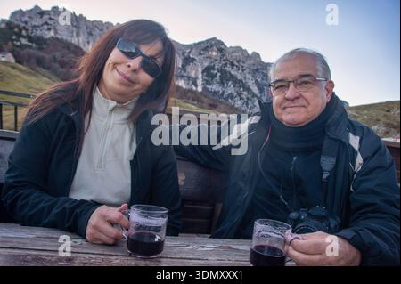 Una coppia più anziana gode di un momento di relax all'aperto, sorseggiando del VIN brulé caldo da tazze di vetro in un paesaggio montano panoramico. Entrambi sono vestiti in inverno j Foto Stock