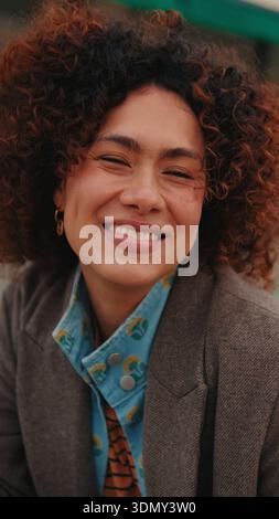 Un ritratto caldo e sincero di una donna sorridente con capelli ricci, con un blazer e una camicia a motivi geometrici. Atmosfera accogliente e sicura, adatta a lavoro e vita Foto Stock