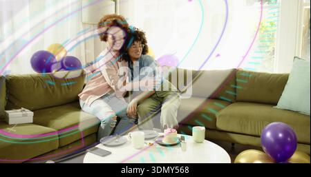 Abbracciando due donne in denim e rosa, sedute su un divano verde oliva a casa con una torta illuminata Foto Stock