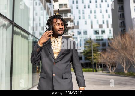Uomo d'affari afroamericano con dreadlock che indossano un abito grigio e una camicia beige, che parla su uno smartphone. Sta camminando fuori di fronte a un moderno Foto Stock