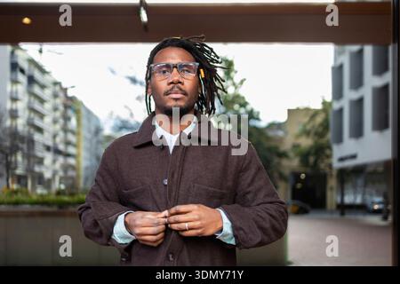 Un fiducioso uomo d'affari afroamericano con dreadlock si erge all'aperto in un ambiente urbano, indossando occhiali e un cappotto, che incarna uno stile moderno A. Foto Stock