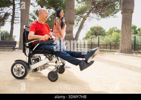 Un uomo felice su una sedia a rotelle ama stare all'aperto con un amico. L'ambiente è un parco illuminato dal sole, che enfatizza l'inclusività, l'amicizia e la gioia dell'essere Foto Stock