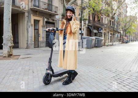 Una giovane donna che indossa un casco e un cappotto beige parla al telefono mentre si trova accanto a uno scooter elettrico in una strada urbana, mescolando moderni mobili Foto Stock