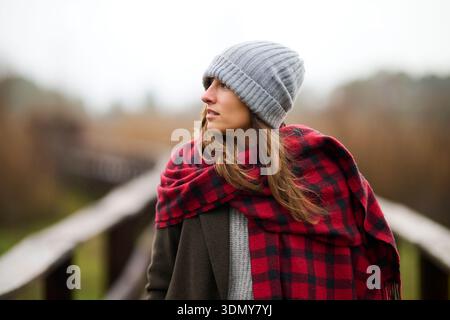 Una donna con i capelli biondi sta all'aperto in una giornata di pioggia, avvolta da una calda sciarpa coperta a quadri e un cappello a maglia. Le piace il bel tempo in mezzo a un s. Foto Stock