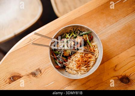 Una ciotola di tagliatelle asiatiche guarnite con verdure fresche e tofu, servita con bacchette su un rustico tavolo di legno. Una rappresentazione perfetta del tradizionale Foto Stock