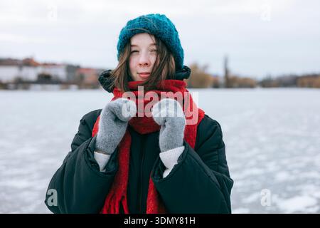 Una donna si stende vestita calorosamente da un lago ghiacciato, indossando un cappello a maglia blu, una sciarpa rossa e guanti. Il paesaggio invernale aggiunge uno sfondo sereno Foto Stock