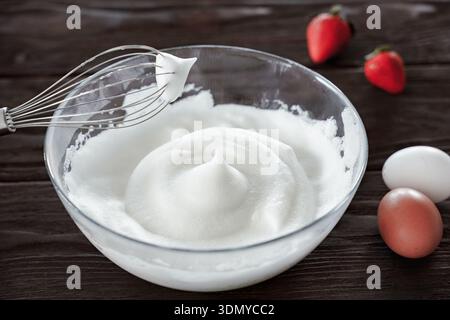 La frusta a mano mescola gli albumi in schiuma spessa, creando picchi stabili nel recipiente trasparente sul piano di lavoro in legno scuro, sul processo di cottura e cottura Foto Stock