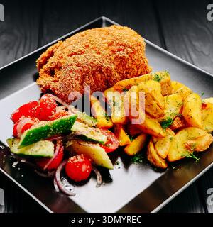 Cotoletta di pollo impanata con patate arrosto e insalata fresca su piatto nero, tavolo scuro Foto Stock