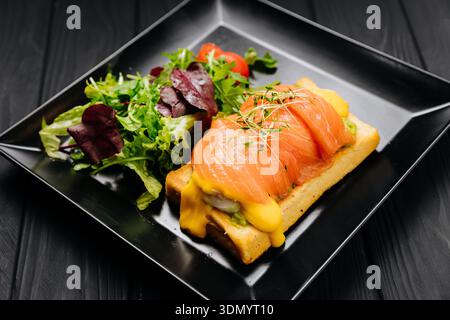 Uova di salmone toast alla benedict con hollandaise e insalata verde su piatto nero, tavolo scuro Foto Stock