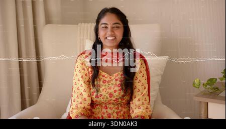 Donna sorridente seduta su una poltrona color crema a casa, indossa un abito giallo e una sciarpa rossa, bindi Foto Stock