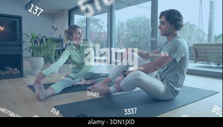 Coppia di stretching che praticano yoga seduto sui tappetini a casa, indossando abbigliamento sportivo con sovrapposizioni per smartwatch Foto Stock
