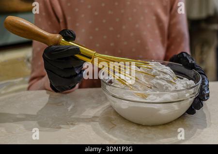Uno scatto ravvicinato di una persona che indossa guanti neri in nitrile utilizzando una frusta dorata di prima qualità per montare una crema bianca o una meringa spessa in un recipiente di vetro Foto Stock