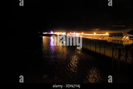 Le luci luminose di un molo costiero brillano nell'oscurità e si riflettono attraverso il mare in una notte buia a Llandudno, nel Galles del Nord. Foto Stock