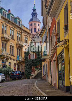 Vista panoramica di una stretta strada acciottolata nella città vecchia di Baden-Baden, Germania, con edifici storici colorati e la guglia della Stiftskirche Foto Stock
