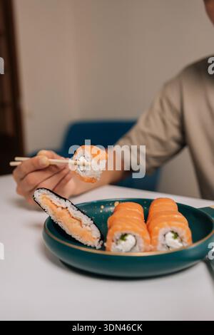 Colpo ritagliato verticale di un uomo irriconoscibile con le bacchette per raccogliere il philadelphia roll dal piatto verde scuro di maki di salmone e il rotolo di riso tagliato Foto Stock
