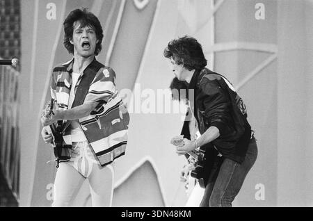 Mick Jagger dei Rolling Stones si esibisce energicamente sul palco, cantando in un microfono e tenendo in mano una chitarra, durante un concerto degli anni '1980 Accanto a lui, Keith Richards (o Ron Wood) suona la chitarra, contribuendo all'iconico sound rock della band. Questa fotografia vintage in bianco e nero cattura l'energia pura di una performance dal vivo. Foto Stock