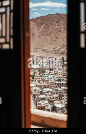 Vista panoramica ad alto angolo della città di Leh e degli edifici della città, vista dalla finestra dell'antico monastero di Tsemo Gompa incorniciata da una tradizionale cornice in legno Foto Stock
