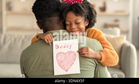 Il bambino abbraccia il padre mentre tiene in mano una carta con il cuore e il papà scritti su di essa in un accogliente soggiorno Foto Stock
