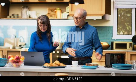 Una coppia di sposi senior gode di un momento tranquillo in cucina con tazze da caffè, guardando un laptop e controllando i siti web. Tranquilla vita in pensione, stile di vita positivo e il piacere della routine. Telecamera B.. Foto Stock
