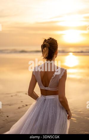 Vista posteriore di una donna in abito bianco che cammina sulla spiaggia sabbiosa e guarda all'oceano durante il tramonto. Momento di pace e romantico concetto di riflessione sul mare. Foto Stock
