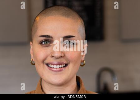 Donna sorridente sui vent'anni con testa rasata e gioielli al bancone del bar con menu lavagna Foto Stock