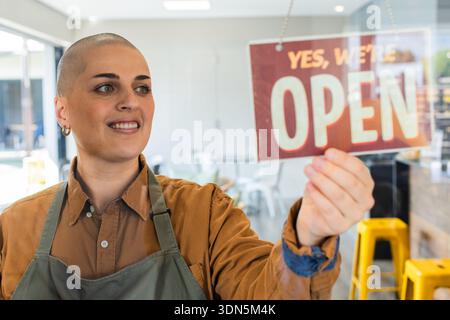 Donna che diventa rossa e bianca con un cartello APERTO su una porta di vetro all'ingresso del caffè che indossa un grembiule verde Foto Stock