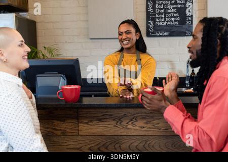 Barista asiatico/indiano di medie dimensioni che serve tazze rosse a due clienti al banco di legno del caffè Foto Stock