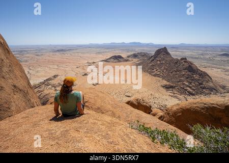 Donna seduta sulle rocce e godendo della vista sullo arido paesaggio desertico di Spitzkoppe, Spitzkoppe, Erongo, Namibia Foto Stock