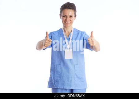Un'infermiera donna positiva in scrub medici blu dà due pollici in su, simboleggiando l'approvazione della salute, il recupero di successo, e la soddisfazione professionale. Foto Stock