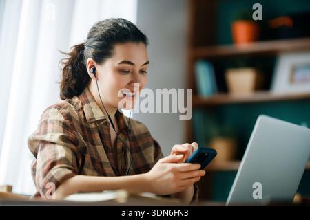 Una donna allegra che indossa le cuffie si impegna a lavorare in remoto da casa, utilizzando smartphone e laptop. La configurazione significa tecnologia moderna, connetti Foto Stock