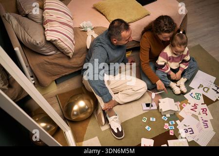 Uomo di mezza età caucasico, donna adulta caucasica e bambina caucasica seduta sul pavimento impegnata in attività educative con schede alfabetiche e materiali di apprendimento Foto Stock
