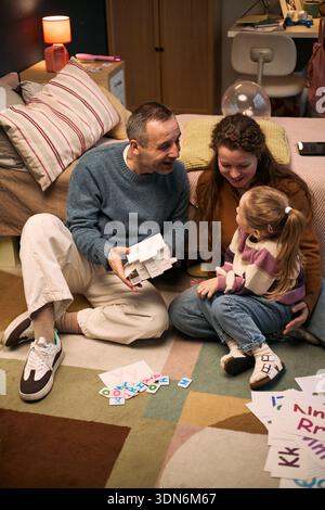 Uomo di mezza età caucasico e donna caucasica seduti sul pavimento che si impegnano con una bambina caucasica, che tiene una casa modello e gioca insieme a un gioco educativo all'interno Foto Stock