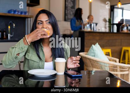 Donna asiatica indiana che morde pasticceria e tiene una tazza di caffè al tavolino nero lucido con smartphone Foto Stock