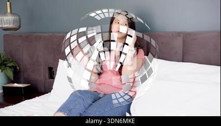 Donna asiatica parlante che tiene lo smartphone all'orecchio sul letto, indossa T-shirt rosa e jeans Foto Stock