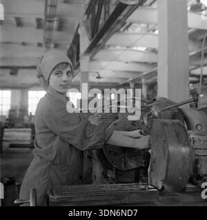 Sloviansk, regione di Donetsk, Ucraina, URSS. 1960 anni Fotografia storica d'archivio. Una giovane donna che lavora su un tornio nell'officina meccanica della mietitrebbia ceramica Sloviansk. Indossa un cappotto da lavoro protettivo e un velo durante il funzionamento di attrezzature industriali pesanti. La donna guarda con fiducia la telecamera. Grandi finestre di fabbrica e pilastri in cemento sono visibili sullo sfondo sfocato. Questa autentica immagine in bianco e nero cattura il lavoro tecnico qualificato delle donne sovietiche nella pacifica regione del Donbas Foto Stock