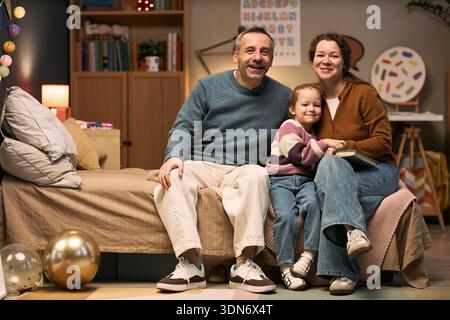 Uomo caucasico di mezza età, donna e bambino caucasico seduti insieme sul letto sorridente, bambino abbracciato donna, famiglia che passa il tempo insieme in un ambiente accogliente Foto Stock
