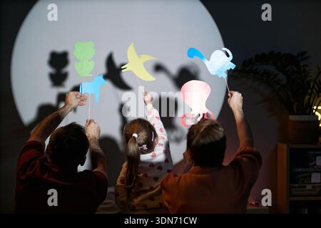 Uomo caucasico, donna caucasica e bambino caucasico che tiene in mano marionette di carta colorata di fronte al muro illuminato, creando forme animali con le mani alzate insieme Foto Stock