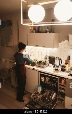 Donna che lava i piatti in una cucina moderna con lavastoviglie a vista. La cucina ha un design elegante, con piastrelle esagonali e illuminazione calda. Vari ki Foto Stock