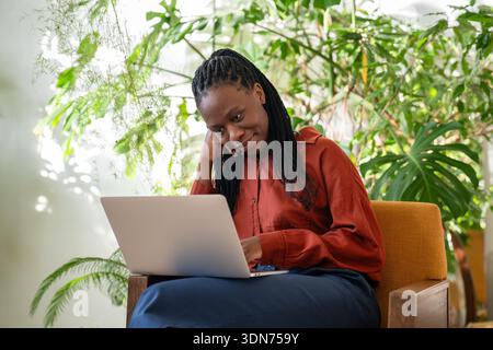Una donna d'affari afro-americana rilassata e sorridente lavora con un computer portatile in un accogliente giardino tropicale. Foto Stock