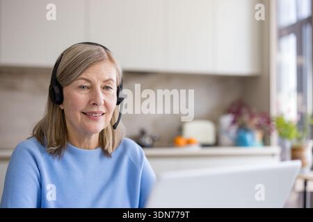 Donna anziana con auricolare sorridente durante una videochiamata con un notebook dalla sua cucina di casa, lavora da remoto come consulente esperto di tecnologia che si connette Foto Stock