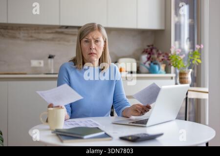 Donna anziana a casa che detiene documenti e gestisce le finanze personali, sentendosi frustrato e stressata per tasse, debiti e aumento dei costi, calcul Foto Stock