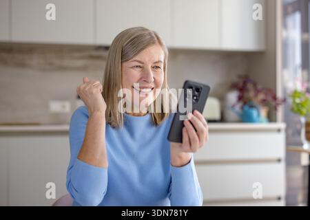 Donna anziana felice che vive un momento di successo, guardando lo schermo di uno smartphone con un'espressione gioiosa e alzando il pugno in trionfo mentre ci si trova Foto Stock